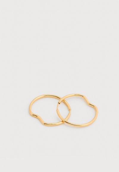 Δύο λεπτά, λαμπερά χρυσά σκουλαρίκια κρίκοι με διακριτικές οργανικές κυματιστές μορφές, τοποθετημένα επικαλυπτόμενα σε απλό ανοικτό φόντο.