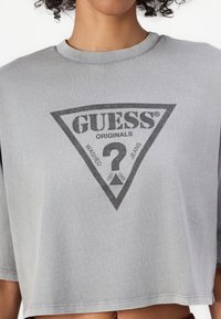 Grå, kort T-skjorte laget av bomull med rund hals og korte ermer. Har et svart trekantet logo med teksten "GUESS ORIGINALS" og spørsmålstegn.