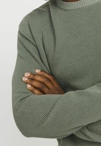 Grön texturerad stickad tröja med rundad halsringning. Har en ribbad krage och långa ärmar med en avslappnad passform, som visar ett våffelmönster.