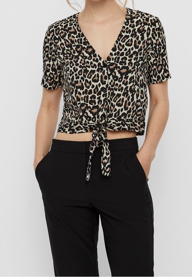Femme portant un chemisier court à manches courtes imprimé léopard noué et un pantalon noir taille haute, mains dans les poches, cadré au niveau du ventre.