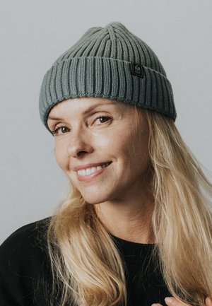 Glimlachende vrouw met lang blond haar, een grijze ribgebreide muts en een zwart topje, tegen een lichte effen achtergrond.