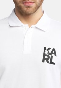 Polo bianco con colletto strutturato, patta con due bottoni e un grande logo grafico nero sul lato sinistro del petto.