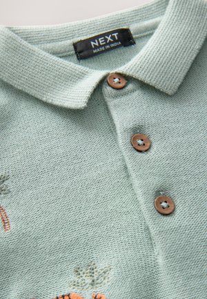 Polo in tessuto a maglia verde chiaro con colletto appuntito, tre bottoni marroni e sottili dettagli ricamati. Etichetta del marchio visibile.