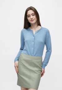 Cardigan azul claro com botões, mangas compridas, feito de tecido macio. Combina com uma mini saia de couro verde com textura lisa.