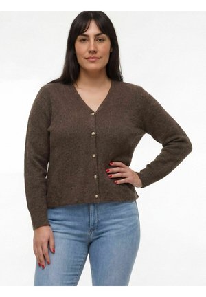 Vrouw met lang donker haar draagt een bruine vest met knopen en lichtblauwe spijkerbroek, staand met één hand op haar heup tegen een witte achtergrond.