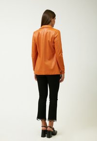 Blazer de imitación cuero naranja con textura suave, mangas largas y un corte entallado. Combinado con pantalones negros con bordado de encaje en los dobladillos.