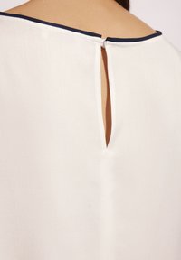 Biała bluzka z tkaniny z okrągłym dekoltem wykończonym granatowym obszyciem oraz otworem w szwie z tyłu. Gładka faktura i prosty krój.