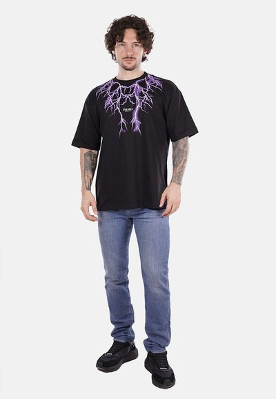 T-shirt nera con grafica a fulmine viola e logo, abbinata a jeans azzurri chiari e sneaker nere. Il modello ha i capelli ricci e dei tatuaggi.