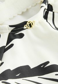 Schwarz-weißer Stoff mit floralem Muster zeigt einen goldenen Verschluss mit rechteckigem Detail in der Nähe des Halsausschnitts. Glatte Textur, leichtes Design.