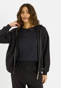 Sweat à capuche zippé noir à coupe décontractée, fabriqué en tissu doux. Comprend une capuche, une fermeture éclair argentée, et est assorti à un haut noir assorti.