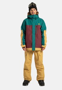 Chaqueta de nieve en turquesa y marrón con acentos amarillos; cuenta con un cierre frontal, bolsillos y una capucha. Combinada con pantalones de nieve amarillos y botas negras.