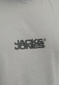 Sivi majica s kratkim rukavima, koja na prsima ima crni, blago zamagljen logo 'JACK & JONES', izrađena od lagane tkanine s glatkom teksturom.