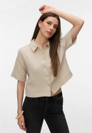 Kobieta o długich brązowych włosach, ubrana w beżową koszulę na krótki rękaw zapinaną na guziki oraz czarne jeansy, podnosząca jedną rękę nad głowę.