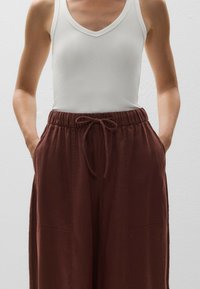 White ribbed tank top with a deep V-neck, paired with loose, rust-colored pants featuring an elastic waistband, drawstring, and side pockets.
