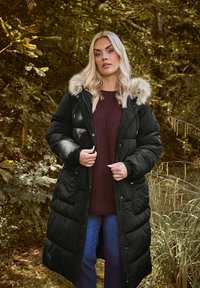 Czarna kurtka puchowa z kapturem wykończonym futrem, z pikowanym wzorem i zapięciem na zatrzask, noszona na teksturowanym burgundowym swetrze.