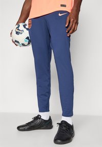 Marineblå Nike joggers med tapered fit, der har et orange logo-patch og åndbart materiale. Sorte sko med grønne accenter og hvide sokker.