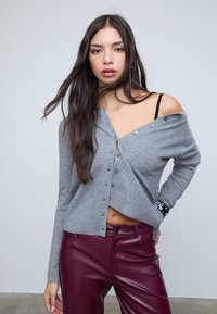 Jonge vrouw met lang donker haar draagt een grijze cardigan met knopen van één schouder en bordeauxrode leren broek, staande tegen een lichte achtergrond.