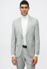 Fato cinza claro sob medida com um padrão de xadrez subtil, usado sobre um suéter de gola alta creme. O conjunto inclui um cinto preto e apresenta dois bolsos frontais.