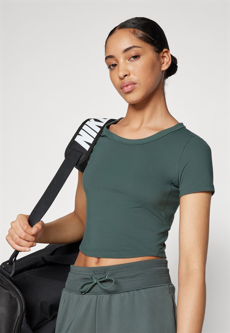 Grünes kurzes T-Shirt mit kurzen Ärmeln aus dehnbarem Material und Rundhalsausschnitt. Das Modell hält eine schwarze Sporttasche mit einem weißen Logo-Trageriemen.