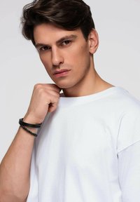 Biała koszula z krótkim rękawem, gładka tekstura. Osoba ma na nadgarstku czarną bransoletkę z koralików. Neutralne tło. Bezpośrednie spojrzenie.