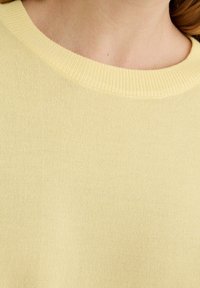 Pull en maille jaune avec un col rond classique, une texture douce et des détails en côtes le long du col. La vue en gros plan met en valeur la qualité du tissu.