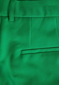Gros plan de pantalon en tissu vert vif montrant la boucle de ceinture, la couture de la taille et le détail horizontal de la poche arrière.