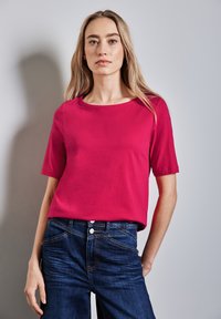 Fuchsiový top s krátkým rukávem vyrobený z měkké tkaniny, s kulatým výstřihem a volným střihem. Doplněno tmavě modrými džínami.