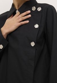 Blusa nera con colletto e bottoni irregolari argentati sul davanti, con una texture liscia e un design strutturato.
