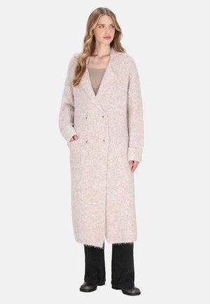 Femeie cu părul lung și ondulat, purtând un cardigan lung din tricot bej, un top taupe, pantaloni negri și pantofi negri, stând cu mâinile în buzunare.