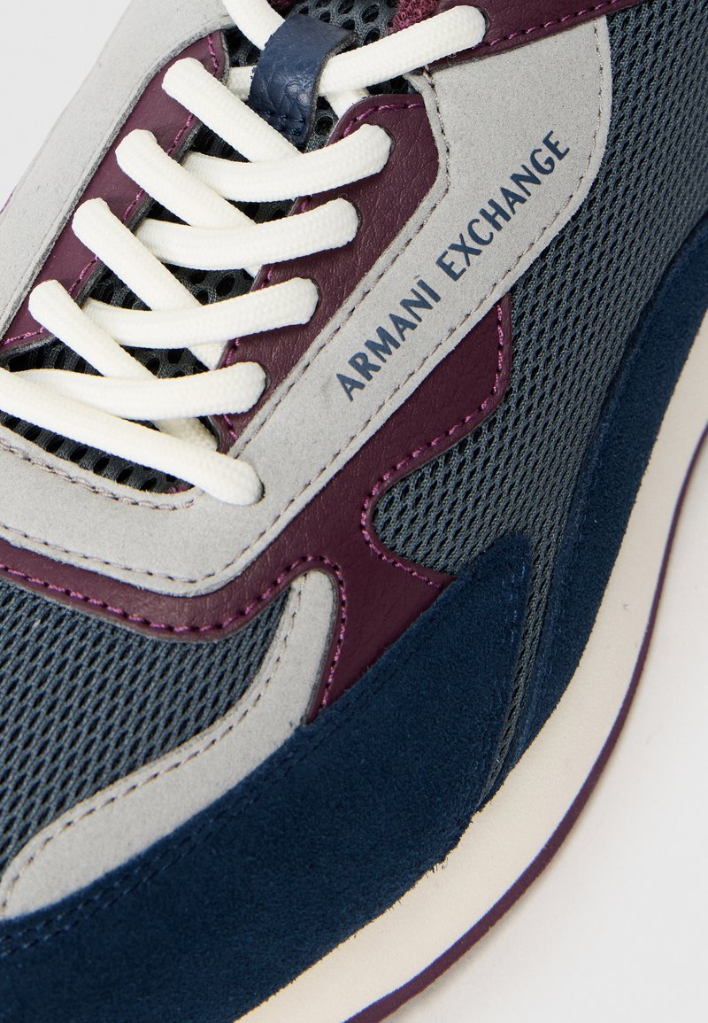 Gros plan sur une sneaker grise, bleu marine et bordeaux avec des lacets blancs et une étiquette "Armani Exchange" sur les panneaux en maille et en daim.