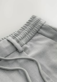Nærbillede af grå sweatpants livrem med sammenkrøllet elastik, bæltestrop og justerbar snor på stofbaggrund.