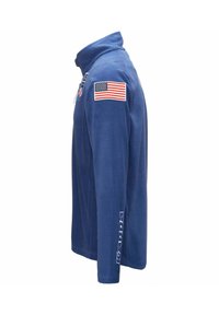 Giacca di fleece blu con colletto alto, caratterizzata da un patch della bandiera americana sulla spalla sinistra e testo bianco "Kaapa" lungo la manica.