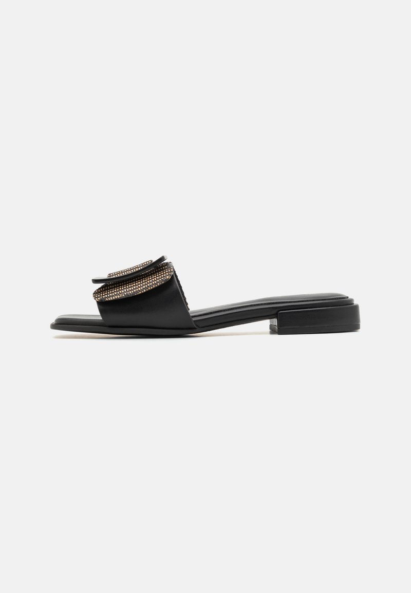 Sandale plate noire à talon bas avec une bride ornée de clous dorés pliée sur le dessus du pied.