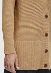 Svetlehnědý žebrovaný cardigan s vertikálními a horizontálními texturovanými vzory, viditelnými hnědými knoflíky a dlouhými rukávy.