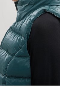 Veste sans manches matelassée bleu sarcelle avec un col montant, présentant un motif matelassé et un tissu brillant et lisse. Portée par-dessus un haut noir à manches longues.
