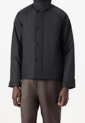 Sort isoleret jakke med høj krave, knaplukning fortil og sidelommer. Har en glat, mat tekstur og en afslappet pasform.