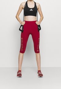 Rote, kurze Leggings mit Ausschnitten an den Seiten, kombiniert mit einem schwarzen Sport-BH. Das Model hält schwarze Fitnessbänder und trägt rote Schuhe.