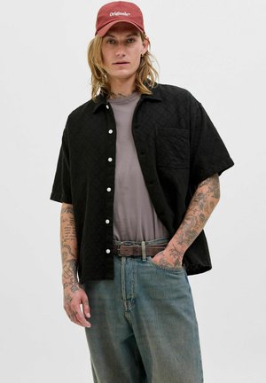 Chemise noire à manches courtes avec un motif texturé et une poche avant, superposée sur un t-shirt gris, assortie à un jean en denim bleu délavé et une casquette rouge.