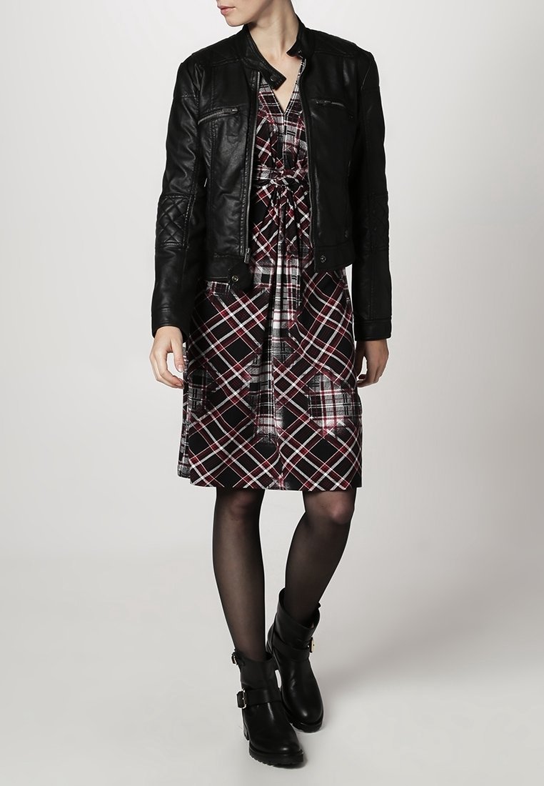 Femme portant une veste en cuir noire, une robe à carreaux avec un nœud à l'avant, des collants noirs transparents et des bottines noires à boucles, debout devant un fond uni.