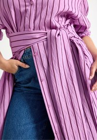 Lyse lilla skjortekjole med lodrette striber, der har bindebånd i taljen, knapper foran og korte ærmer. Bruges over blå denim.