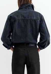 Kobieta ubrana w czarną, krótką kurtkę dżinsową z podwiniętymi rękawami oraz czarne jeansy z wysokim stanem, widoczna od tyłu na białym tle.