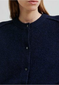 Navy ullcardigan med ribbet rund hals, med svarte knapper og en myk, teksturert overflate. Nærbilde som fokuserer på den øverste delen.