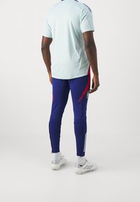 Ljusblå idrottströja med korta ärmar, kombinerad med navyfärgade, figurnära byxor med röda accenter och sidosträck, avslutad med vita fotbollsskor.