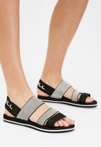 Mustat ja hopeanväriset sandaalit, joissa on kolme säädettävää hihnaa, litteä pohja ja huomaamaton valkoinen raita reunan ympärillä. Teksturoidut kankaat ja joustavat yksityiskohdat.