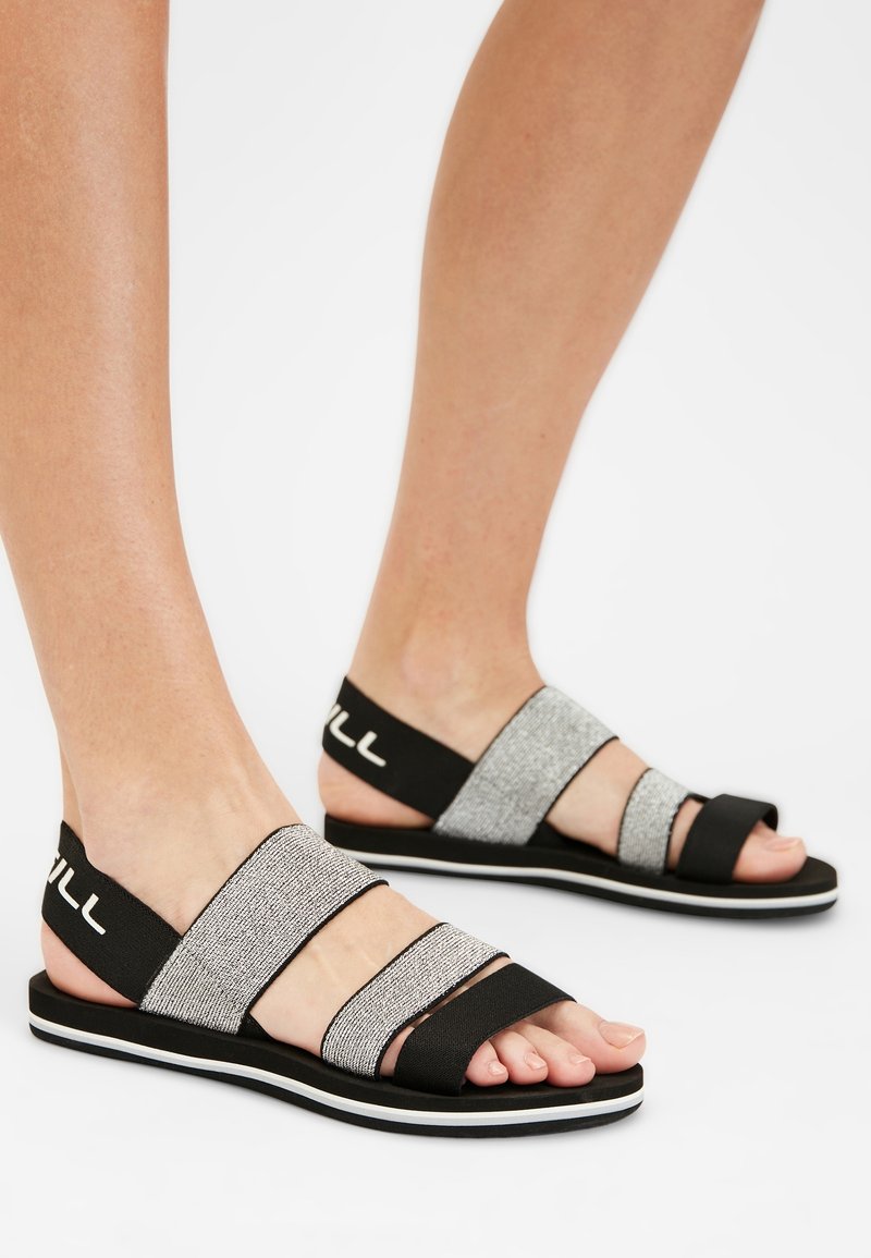 Mustat ja hopeanväriset sandaalit, joissa on kolme säädettävää hihnaa, litteä pohja ja huomaamaton valkoinen raita reunan ympärillä. Teksturoidut kankaat ja joustavat yksityiskohdat.