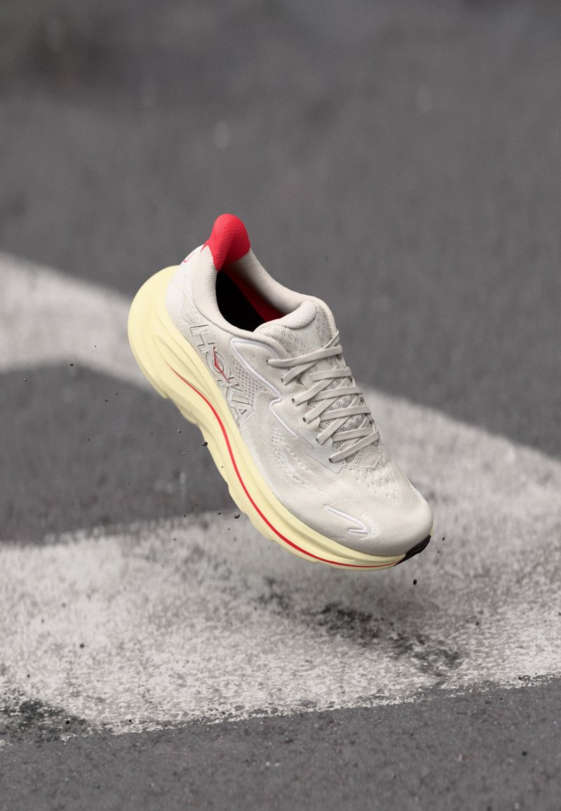 Scarpa da corsa con una tomaia in mesh grigio chiaro, lacci bianchi e una suola gialla con accenti rossi al tallone. Testurizzata per traspirabilità e comfort.
