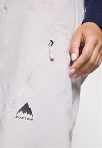 Pantaloni de zăpadă gri deschis, cu un buzunar cu fermoar pe coapsă, având logo-ul negru Burton și o textură de țesătură netedă și durabilă.