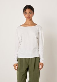 Valge pikkade varrukatega särk, millel on lai kaelus, lõdvestunud istuvus ja pehme tekstuur, mida kombineeritakse oliivroheliste plisseeritud pükstega.