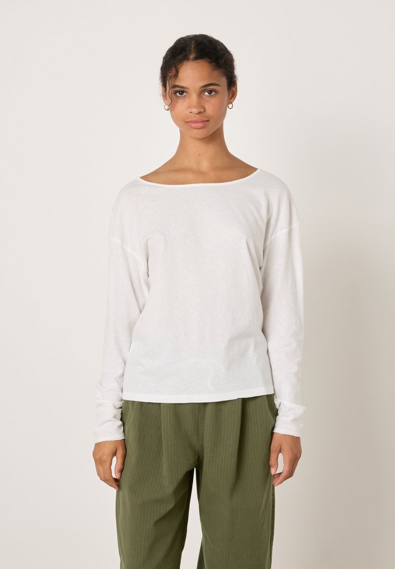 Valge pikkade varrukatega särk, millel on lai kaelus, lõdvestunud istuvus ja pehme tekstuur, mida kombineeritakse oliivroheliste plisseeritud pükstega.