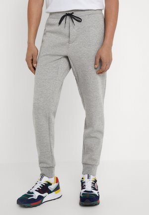 Homme portant un pantalon de jogging gris clair avec un cordon noir et des baskets multicolores, debout sur fond blanc.